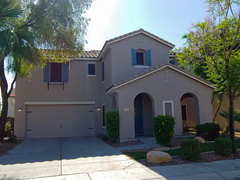 Two-story beige house with a double garage, blue front door, and blue window shutters, surrounded by green shrubs and small trees. The sky is clear and sunny.