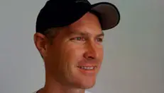 A man wearing a black baseball cap and a white shirt smiles, facing slightly to the right. The background is plain and light-colored.