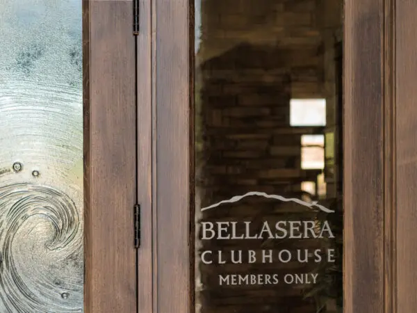 Close-up of a wooden door with a glass panel displaying the text "Bellasera Clubhouse Members Only." A decorative wave-like pattern is etched on the adjacent glass panel.