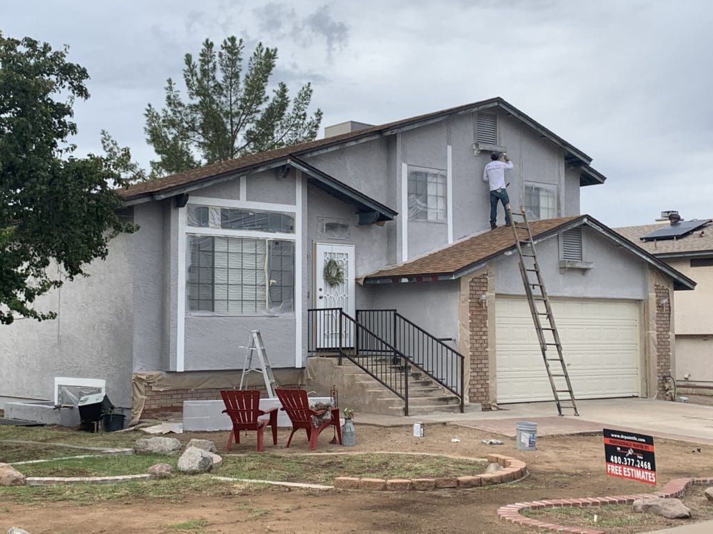 A person stands on a ladder painting the exterior of a two-story house with light gray walls. The front yard has a couple of red chairs, another ladder, and a "Free Estimates" sign near the driveway.