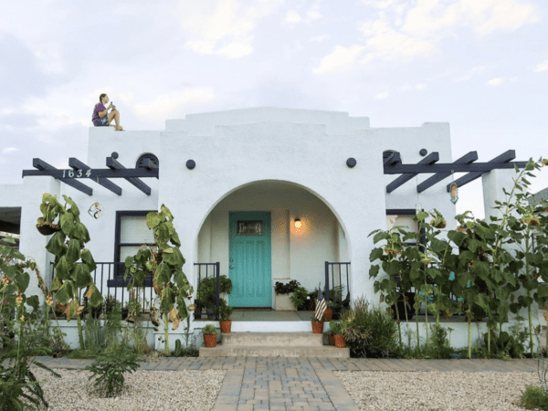 A white stucco house with a teal front door, archways, and potted plants. Sunflowers grow by a gravel path. A person sits on the roof. House number 1034 is visible.