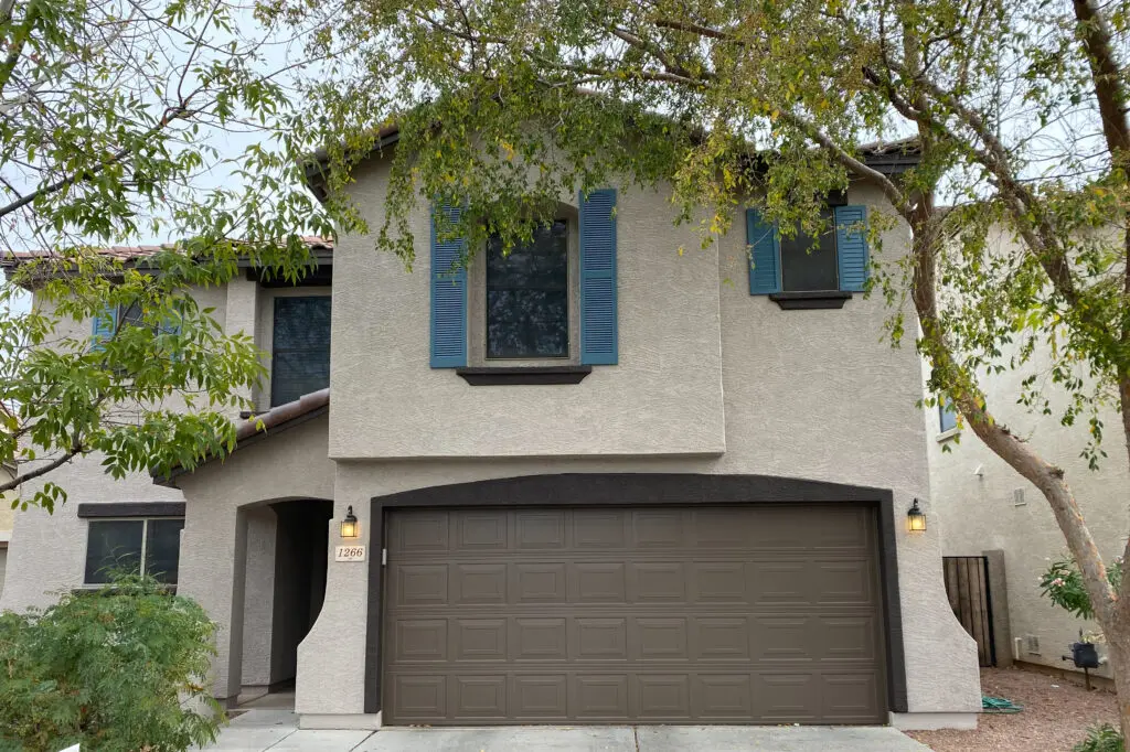 Two-story house with a beige exterior, blue window shutters, and a dark brown garage door. A small tree is positioned in front of the house.