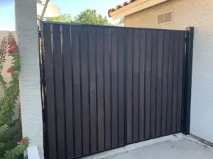 A tall, black wooden fence gate is installed between two sections of a white wall outside a house. A small portion of greenery is visible on the left side.