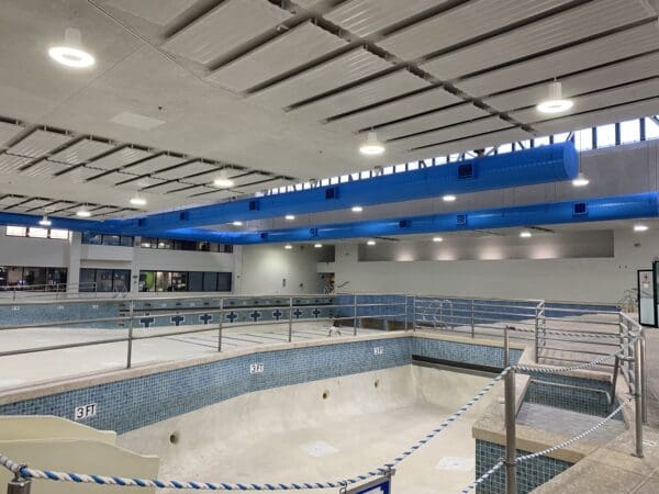 A view of an empty indoor swimming pool under renovation, with blue ventilation ducts on the ceiling and construction barriers around the pool perimeter.