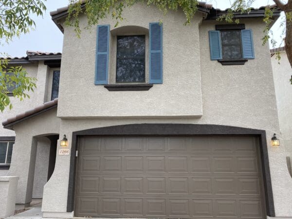 A two-story house with light-colored walls, blue shutters, and a brown double garage door. A small lantern-style light fixture is mounted beside the garage. Tree branches are visible overhead.