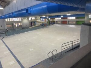 An empty indoor swimming pool area under construction with a large blue ventilation duct overhead and a water slide in the background.