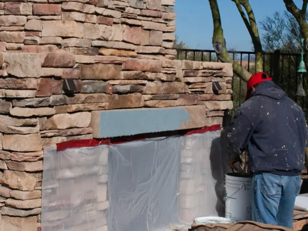 A person in a red cap and blue jacket is working on a stone wall, using supplies from a white bucket. A plastic sheet covers part of the wall. Trees and a fence are in the background.