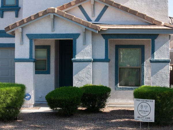 A white house with blue trim has a sign in the front yard that reads "Free Estimates, Dr. Paint." There are neatly trimmed bushes in front and gravel landscaping.