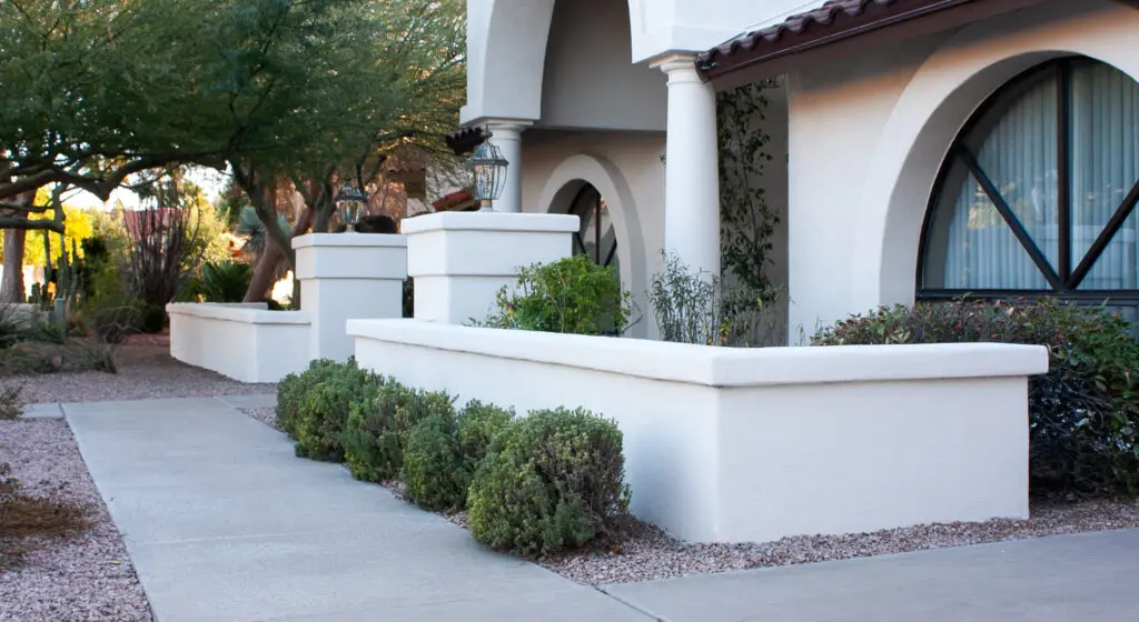 Sidewalk and front view of a white-stucco house with arched windows, bushes, and desert landscaping.