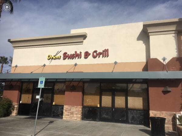 A restaurant storefront with the sign "Sushi & Grill" above the entrance. The facade has a mix of beige and brown colors. There is a "Reserved Parking" sign on a post in front.