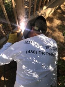 A person wearing a "Dr. Paint" shirt and protective gloves is welding a metal structure outdoors, generating bright sparks. They are surrounded by greenery and wooden slats.