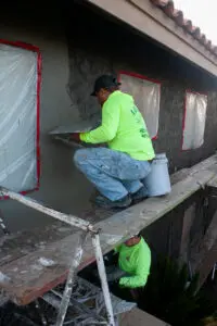 Two construction workers on a scaffold applying stucco to a building's exterior wall, with windows protected by plastic coverings and tape.