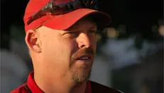 A man with a goatee and sunglasses on his head wears a red cap and red shirt, standing outdoors with a neutral expression.