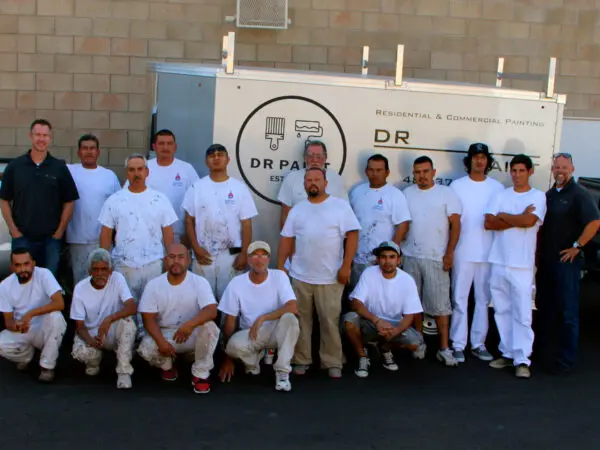 A group of fifteen men posing in front of a truck labeled "DR Paint." Some men are in painting uniforms, while others are in casual clothing. The background features a brick wall.