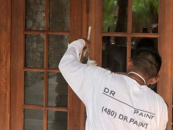 A person with a "DR PAINT" shirt is painting a wooden door with glass panels using a paintbrush.