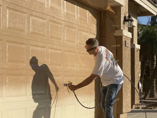 A person in a white shirt and jeans is spray painting a garage door beige, using a paint sprayer. Shadows from the person and the garage door are cast on the surface.