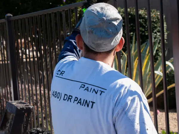 A person wearing a white shirt and a cap is painting a metal fence outdoors. The shirt has text that includes a phone number and "DR PAINT.