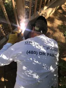 A person wearing a welding helmet and gloves is welding metal outdoors. They are wearing a shirt with text partially visible on the back.